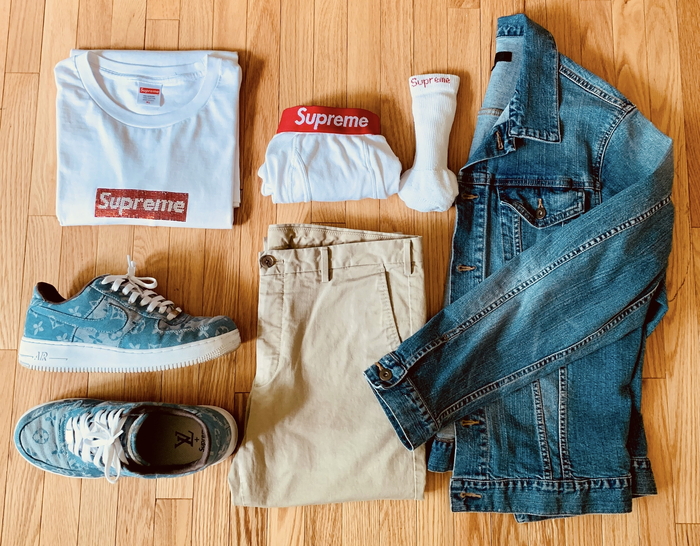 clothes laid out on a wood floor, including a white tee with ‘Supreme' written across, 
                        blue print sneakers, white socks, sand chino trousers and a denim jacket.