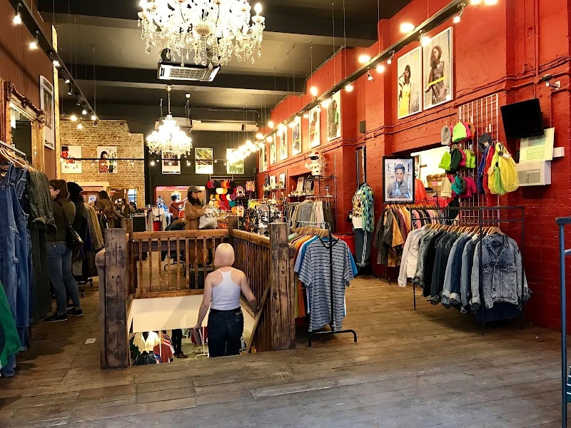 A clothes shop with wooden flooring, chandeliers and red wallpaper. There are pictures on the wall and lots of rails of clothes with people looking around. 
                            A woman in a white vest with a white bob haircut is central to the image, walking down the stairs.