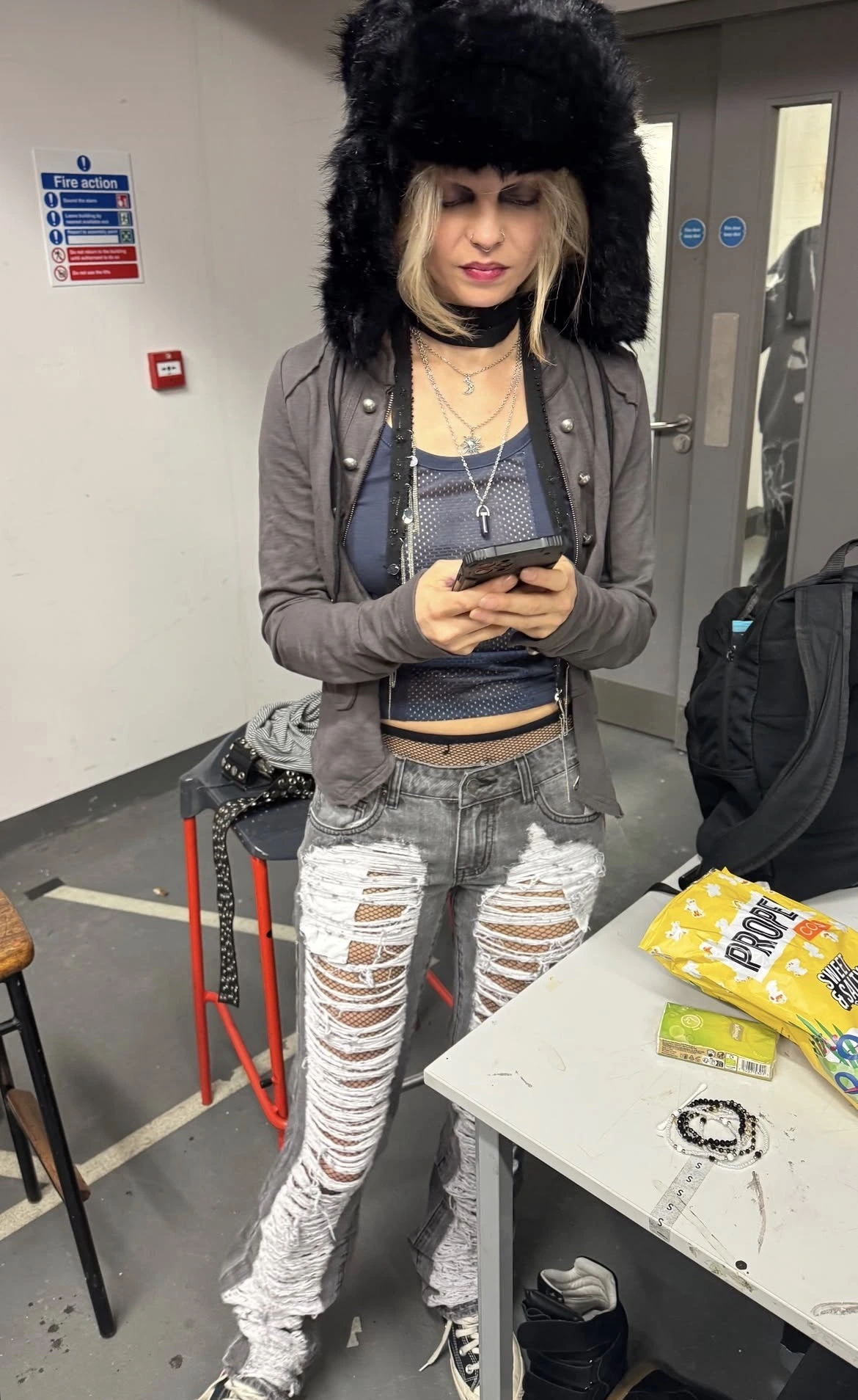 A woman with a black furry hat, wearing a grey jacket and grey and white ripped jeans. She’s standing looking on her phone.