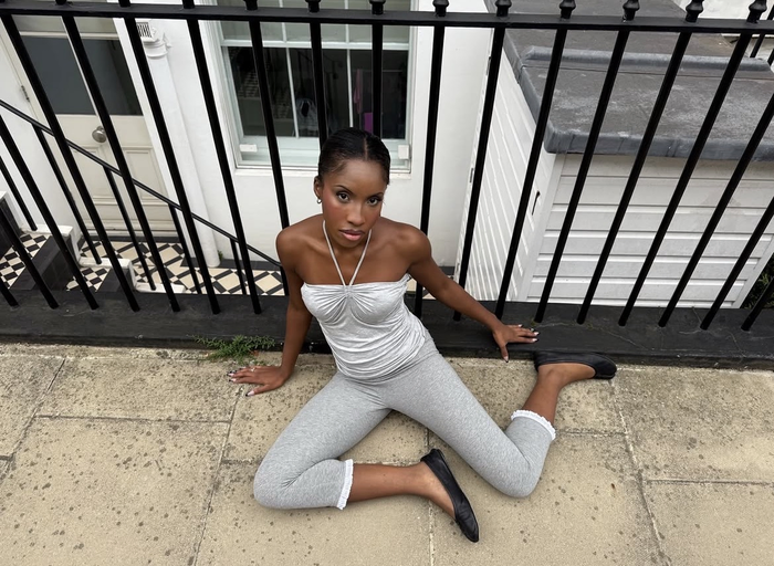 A woman wearing a grey outfit, sat on the floor against black railing and a white house.