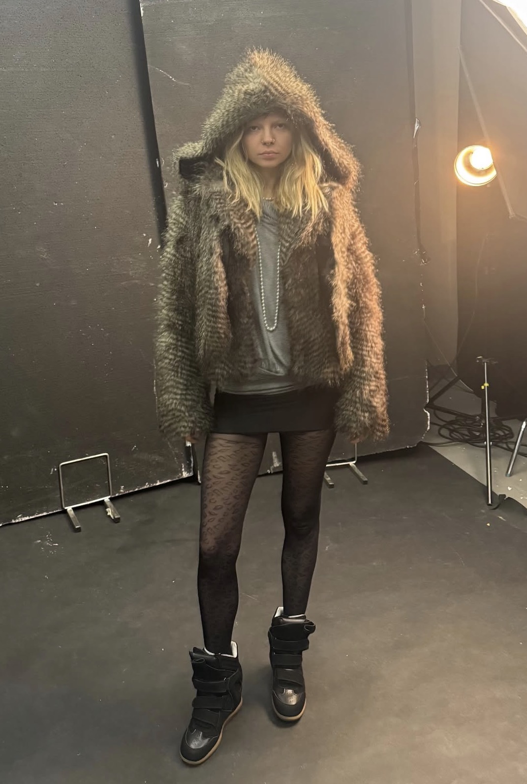 A girl standing against a black background, wearing a furry hooded jacket, a black mini skirt and black ankle boots with patterned tights.