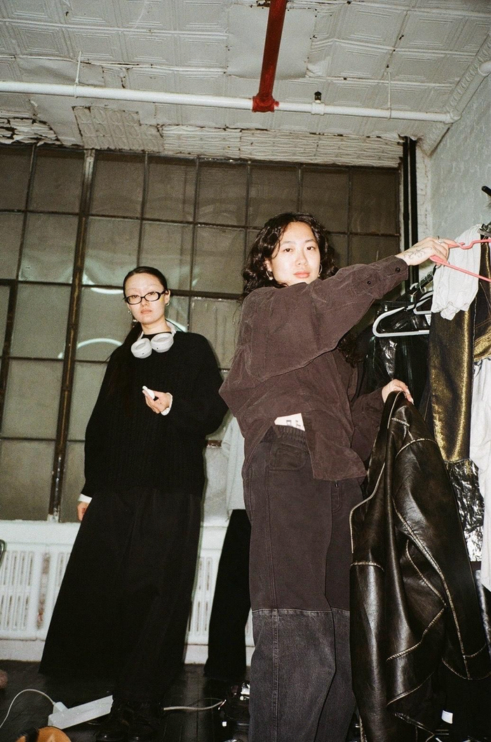 two girls look into the camera inside a room with a white ceiling. The girl to the left wears black. The girl to the right wears brown and holds a coat hanger and clothes.