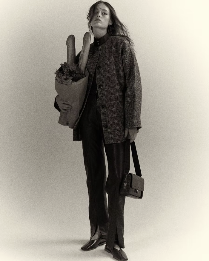 a black and white image of a woman posing infront of a white backdrop. She wears a checked jacket, tailored trousers, and holds a handbag and paper bag with baguettes inside.