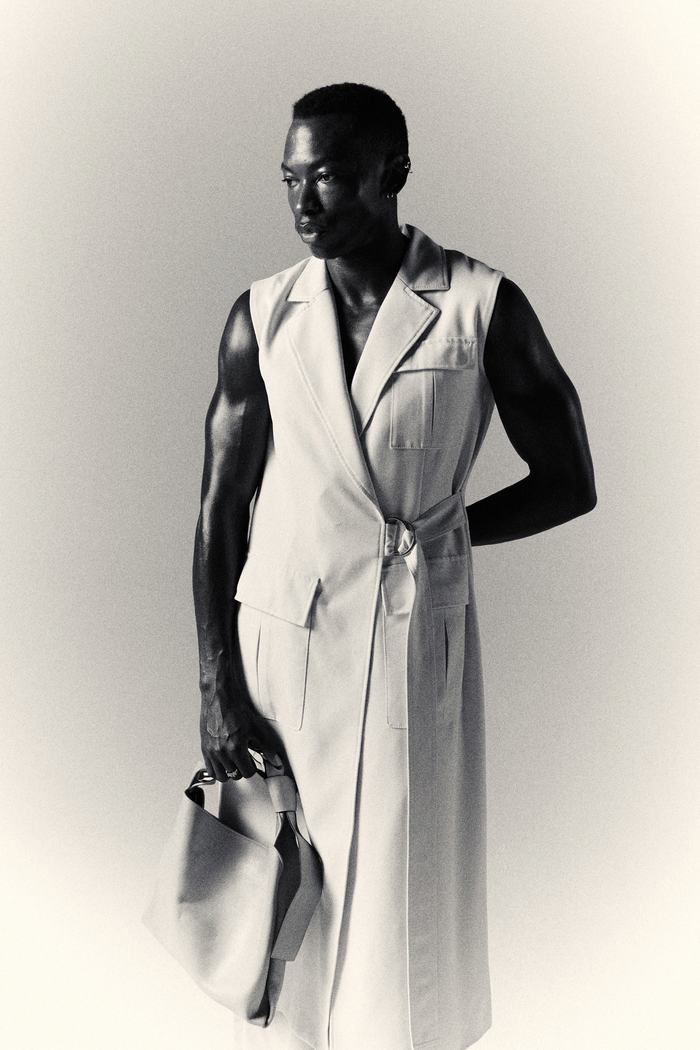 a black and white image of a man posing in a long, white tailored coat and holding a hand bag.