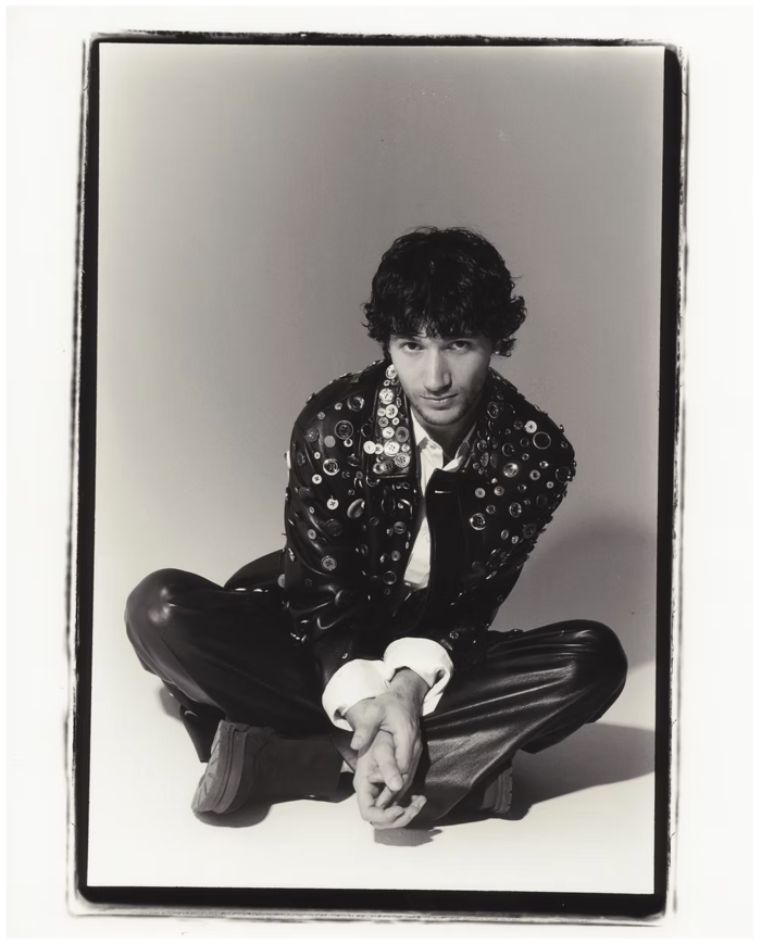 a black and white image of a man sitting crossed-legged, posed with his hands together in front of him and wearing leather trousers and a leather jacket covered in buttons.