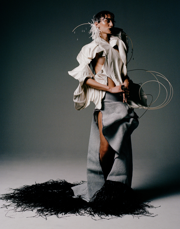 a woman poses against a grey backdrop. She stands on black tassled material. She looks to the right, wearing a white headpiece, a deconstructed corset and long grey skirt.