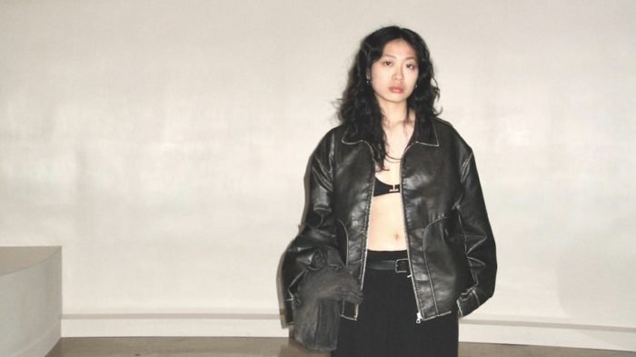 an image of a girl standing against a beige wall, wearing a black leather jacket and black trousers.