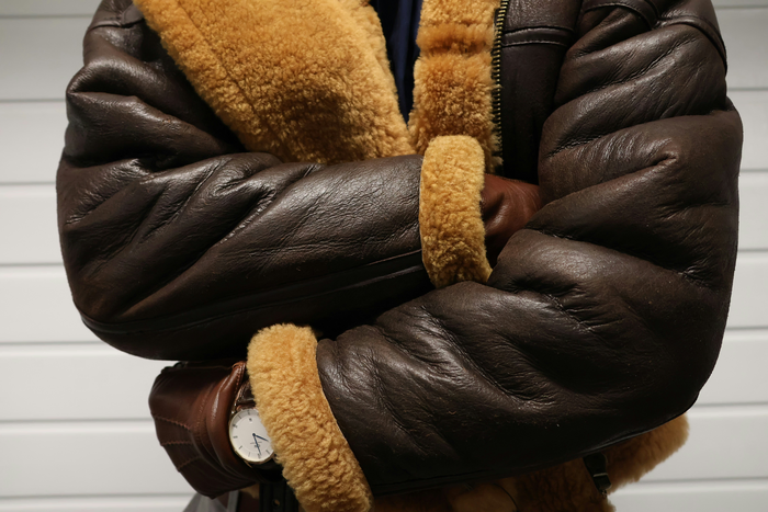 A close up image of a man wearing a brown winter jacket, with fluffy light brown cuffs, with brown gloves and a watch. The background is white.