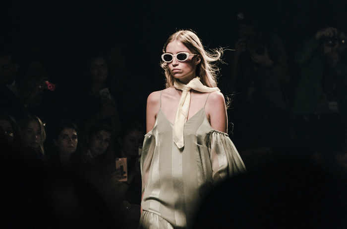 a girl in a pale green dress with sunglasses, a yellow scarf and blonde hair walks down a catwalk.